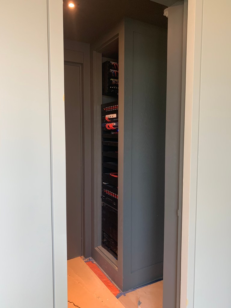 A narrow corridor leading to a cabinet with electronic equipment and wiring, featuring shelves filled with devices, and dim overhead lighting.