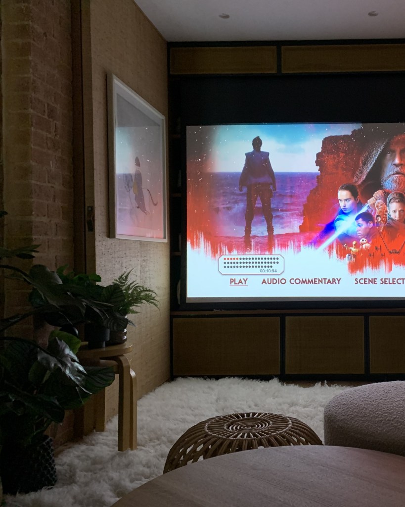 A cozy living room with a projector screen displaying a movie menu, featuring characters and options for playback. There is a circular wooden table and potted plants in the foreground.